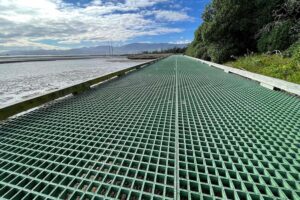 A green FRP grating walkway extends into the distance, flanked by foliage on the right and a body of water on the left under a partly cloudy sky.