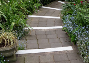 A stone pathway with FRP anti-slip nosing strips is bordered by various green plants and small blue flowers, leading through a garden.