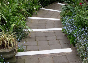 A narrow stone pathway with FRP anti-slip nosing strips on each step is flanked by green foliage and small blue flowers on both sides.