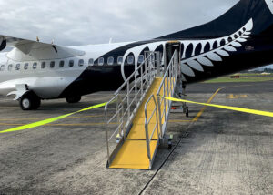 A black and white airplane with a prominent silver fern design on its tail is parked on the tarmac, with a yellow FRP Anti-Slip Sheet mobile boarding ramp leading to the plane's door.