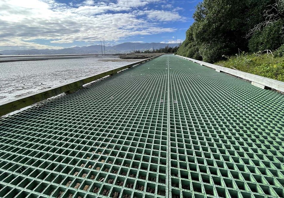 A green FRP grating walkway extends into the distance, flanked by foliage on the right and a body of water on the left under a partly cloudy sky.