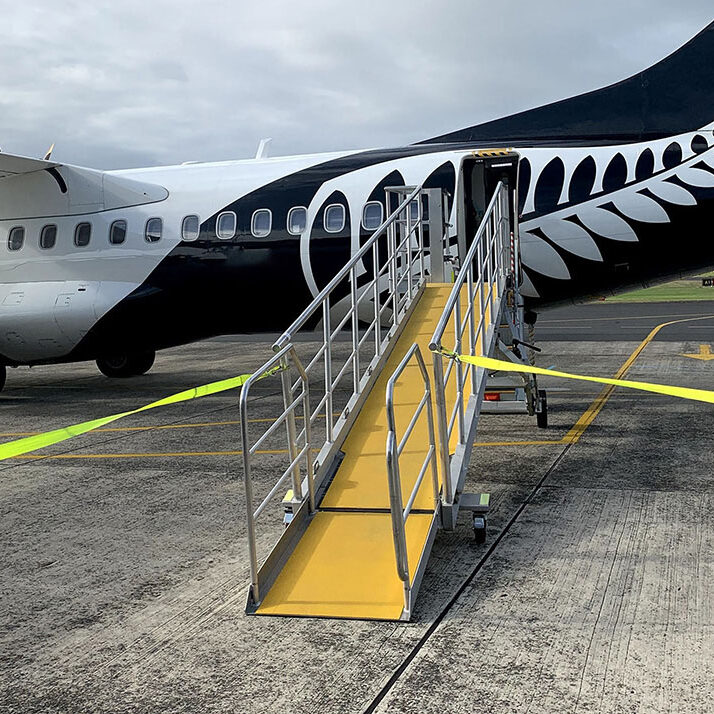 A black and white airplane with a prominent silver fern design on its tail is parked on the tarmac, with a yellow FRP Anti-Slip Sheet mobile boarding ramp leading to the plane's door.