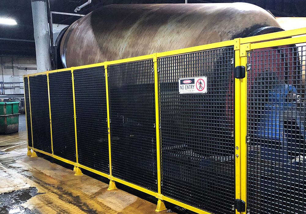 Yellow safety barriers and black FRP modular guarding enclose a large industrial machine. A "No Entry" sign is visible on the fence.