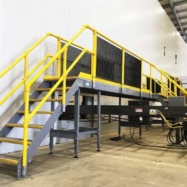 Industrial metal staircase with yellow FRP modular handrails leads up to an elevated platform. Various machinery and cables are visible beneath the platform. The environment appears to be inside a warehouse.